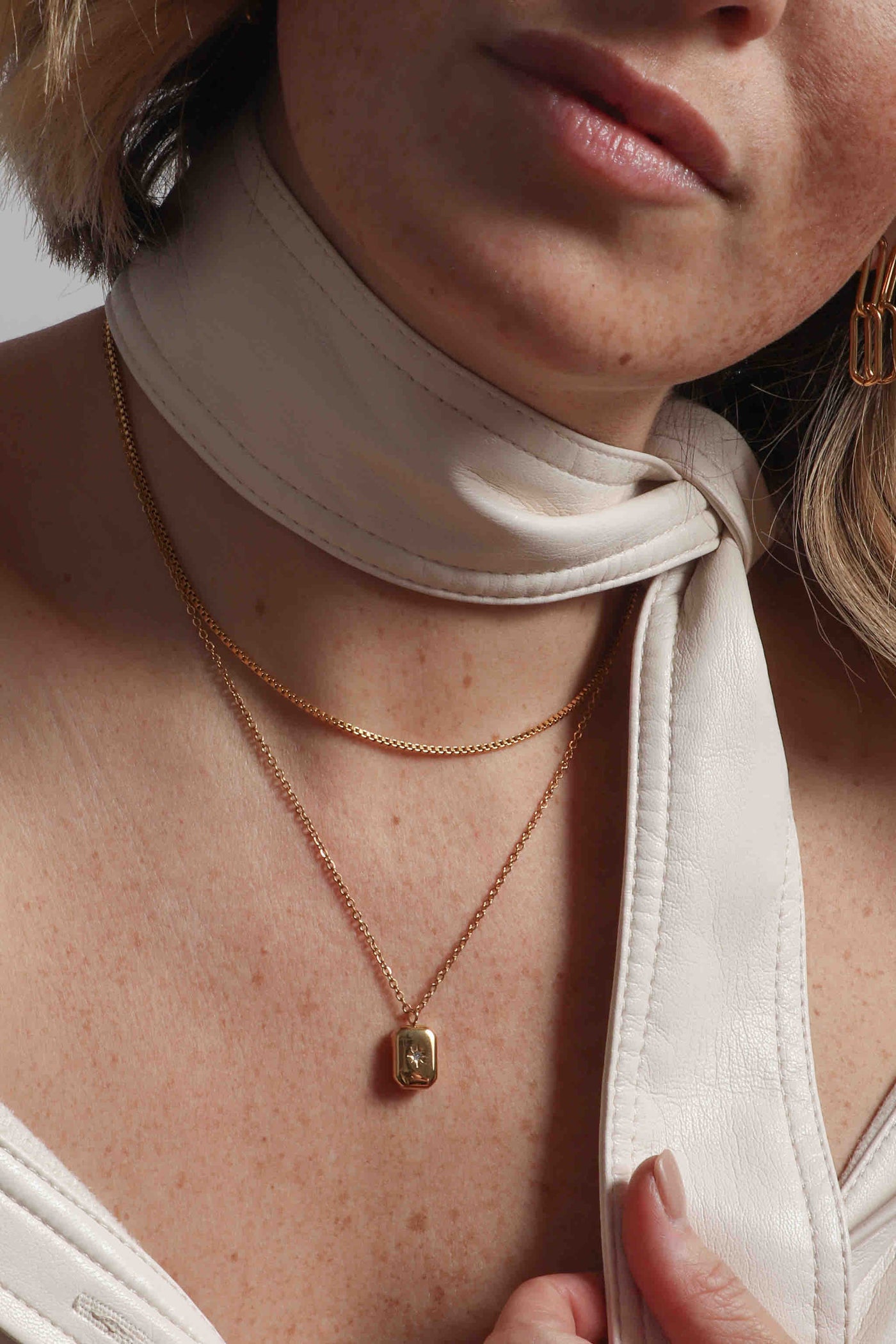 Delicate 14k gold plated stainless steel, hypoallergenic, water resistant, non tarnish, waterproof, rust resistant jewelry by Marrin Costello, featuring our classic paperclip chain Whitney Drop earrings, twisted vintage inspired 5mm rope chain Helix Bracelet, adjustable box chain Nile Choker, and CZ simulated diamond inlaid square signet pendant on a delicate round link chain necklace, styled with a cream vegan leather jacket and neck tie