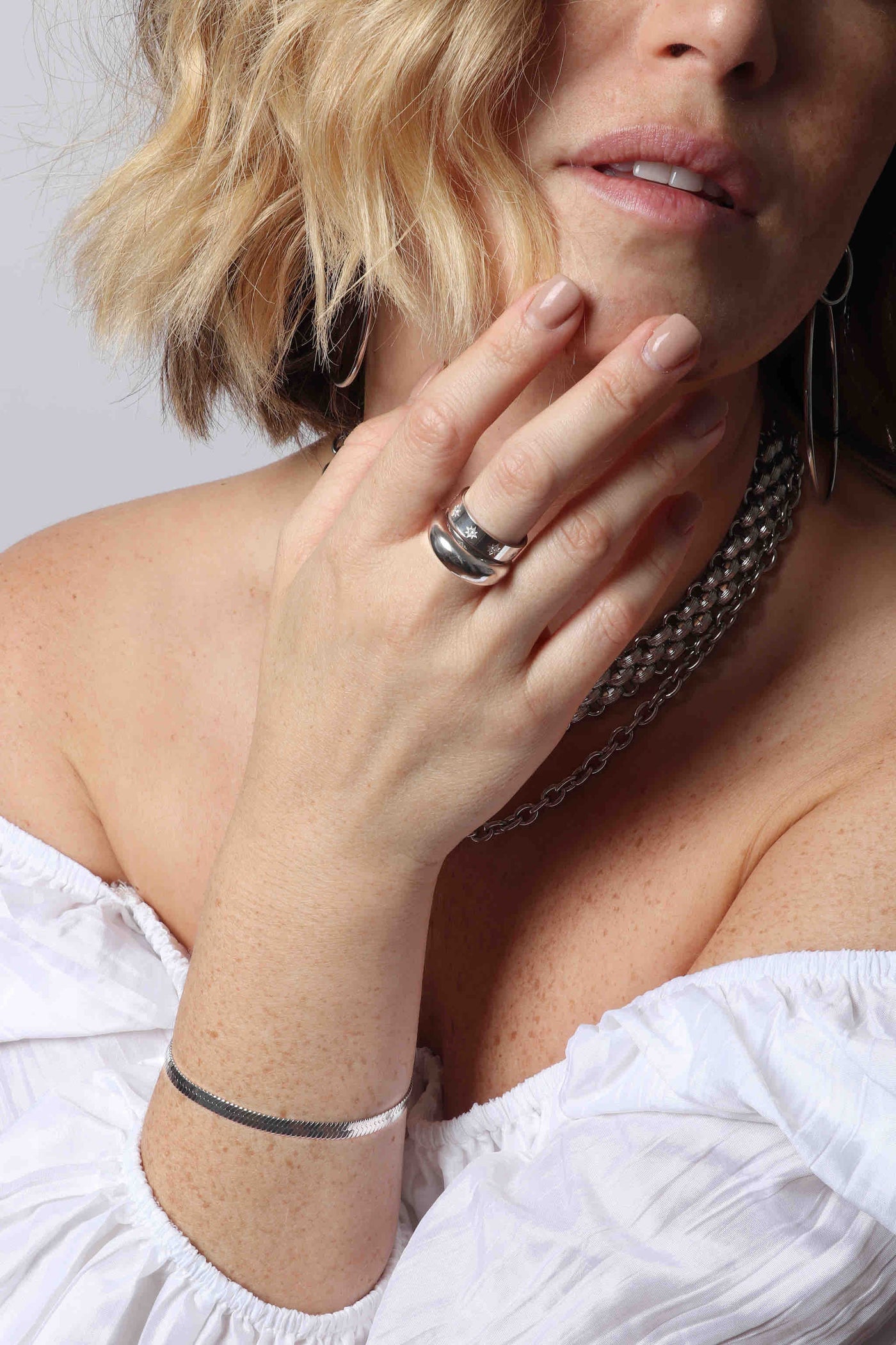 Stacked silver by Marrin Costello Jewelry, including a large 3" double hoop earring, woven textured Lattice XL chain link choker, a smooth polished oval link Mica Chain necklace, dome ring, celestial Cartier inspired ring, and 5mm herringbone Ramsey Bracelet, paired with a white pleated off the shoulder top from Show Me Your Mumu 