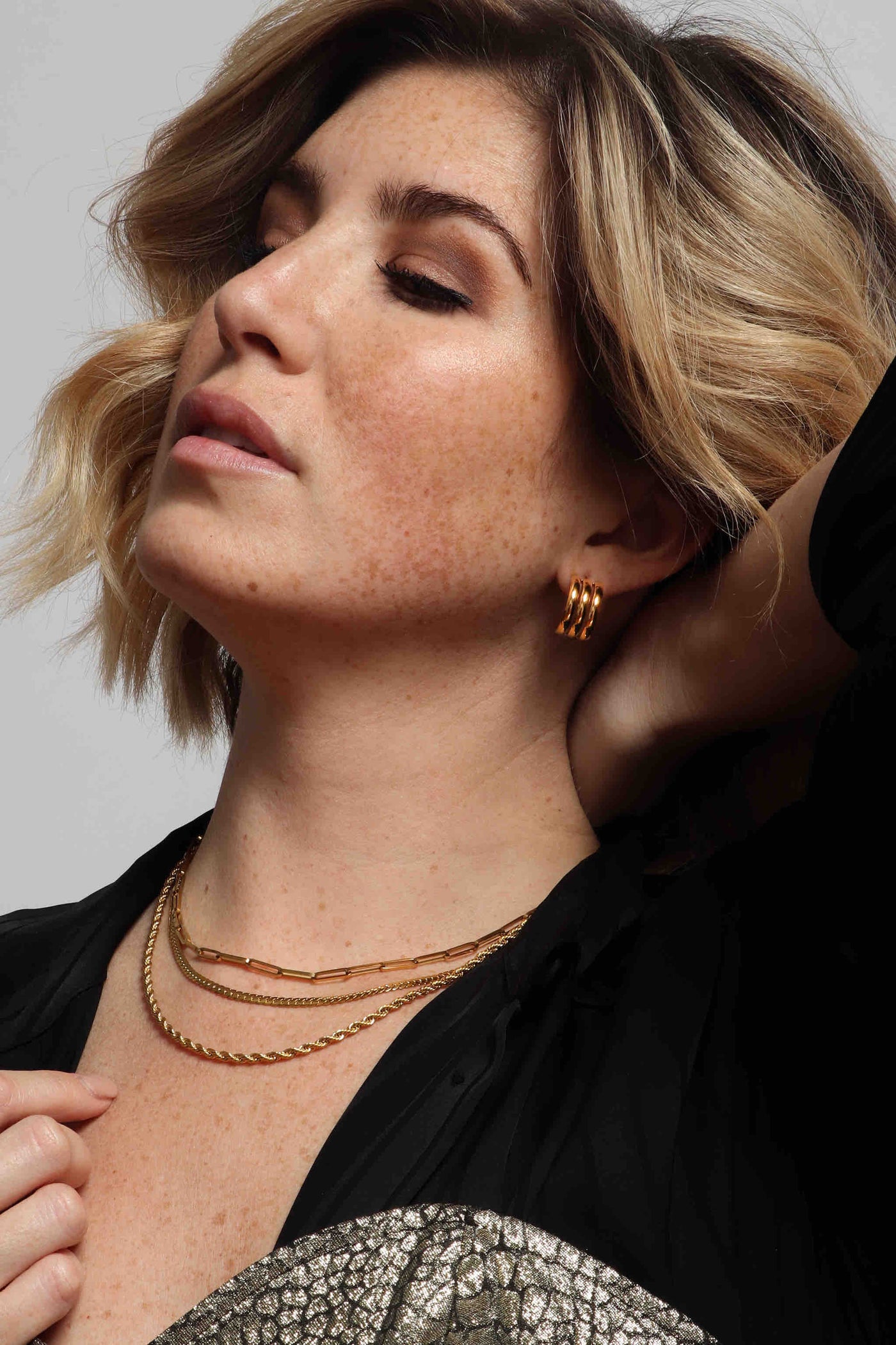 Jewelry stacks by Marrin Costello, featuring our 14k gold plated, stainless steel, water resistant, sustainable layers, like the triple ring Petra hoops, twisted rope Helix Chain in 3mm, flattened Billie Chain in 3mm width, elongated paperclip Empire chain, studded Melrose Band stacker ring, ball chain Crown Band stacker ring, diamond shaped Maverick signet ring, styled with a silk black button down top and metallic alligator embroidered printed bustier top.