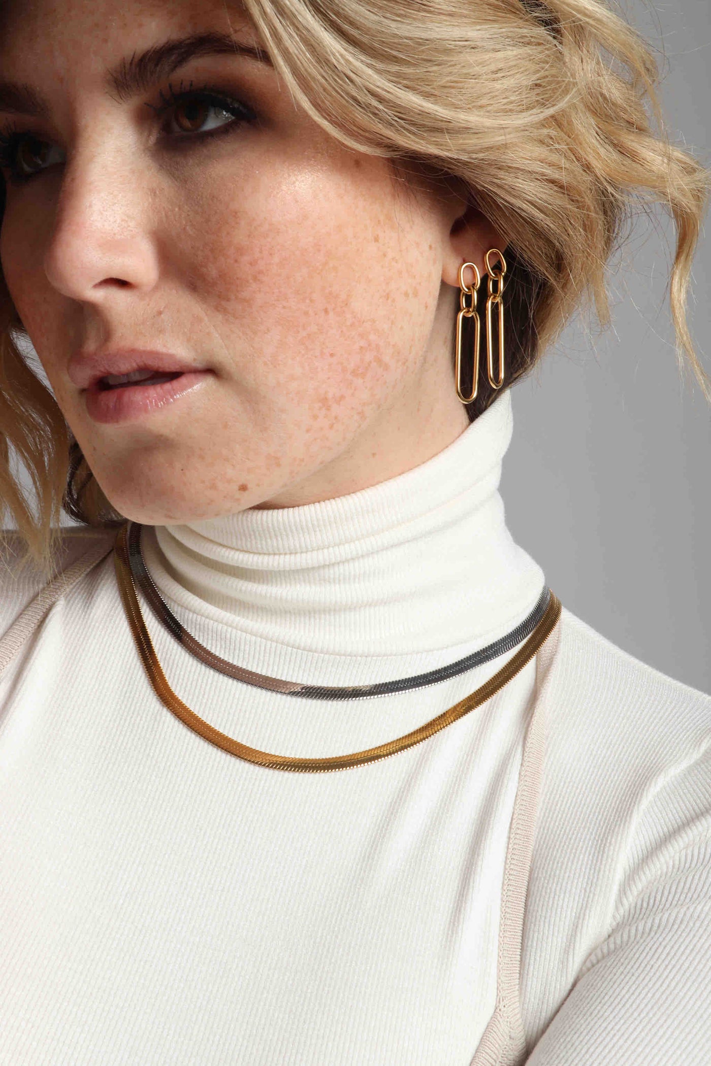 Mixing metals and chain link textures with the Marrin Costello Jewelry collection, featuring the multi oval chain link Mariposa Drops, 5mm vintage inspired classic herringbone Ramsey Chain in mixed metal, all made of polished or 14k gold plated hypoallergenic, water resistant, sustainable, non tarnish stainless steel, styled with a ribbed ivory turtleneck and a mauve ribbed string halter top