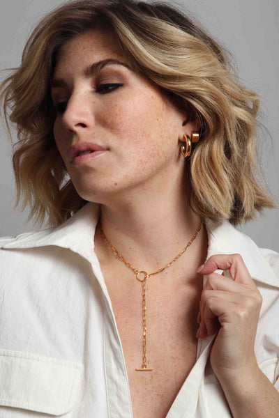 14k gold plated stainless steel, hypoallergenic, water resistant, sustainable, waterproof jewelry by Marrin Costello, featuring our holly everyday Michaela Hoops in 1" diameter, ribbed geometric invisible closure Evelyn 5mm Huggies, and delicate toggle clasp and paperclip link Supreme Lariat necklace, styled with a plunging neckline collared white utility top